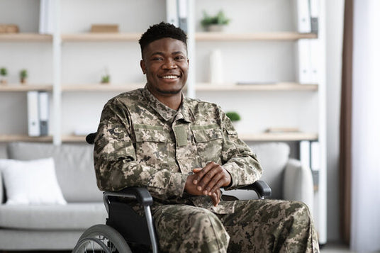 A smiling man in a wheelchair wearing a digital camouflage military uniform, seated in a bright, modern indoor setting with a blurred background.