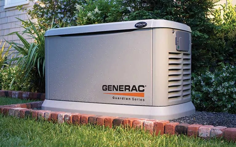A Generac Guardian Series generator sitting outdoors on a brick border in a grassy garden area.