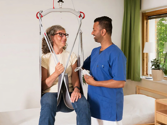 Person using a medical lift with a healthcare provider in a room with a window and plant.