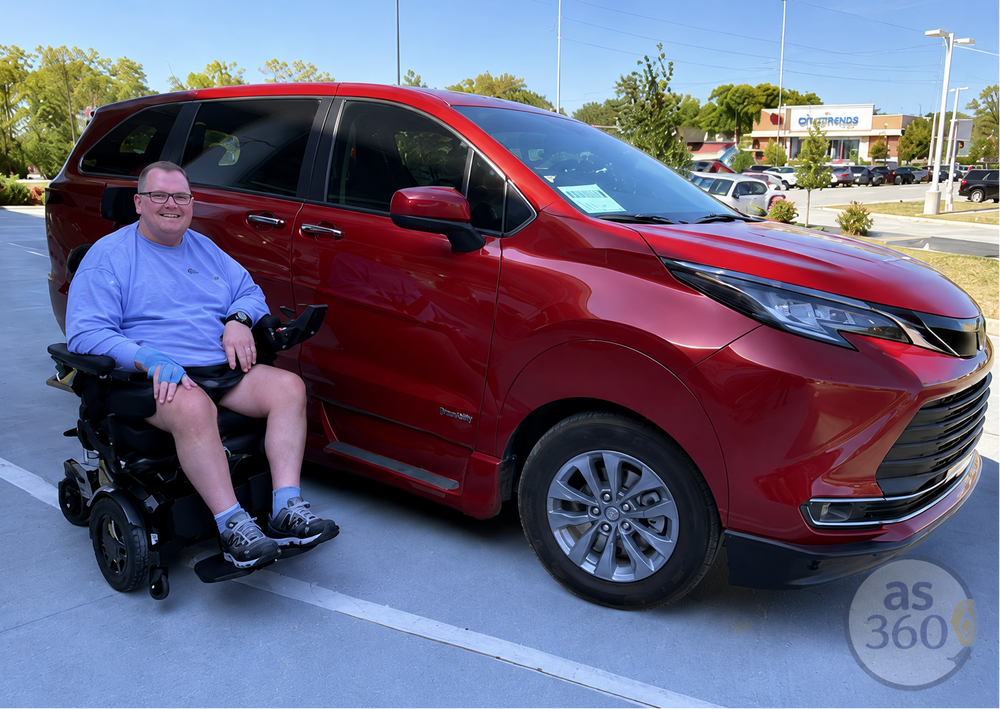 A man in a motorized wheelchair sits beside a red minivan parked in a lot, with trees and a store in the background.