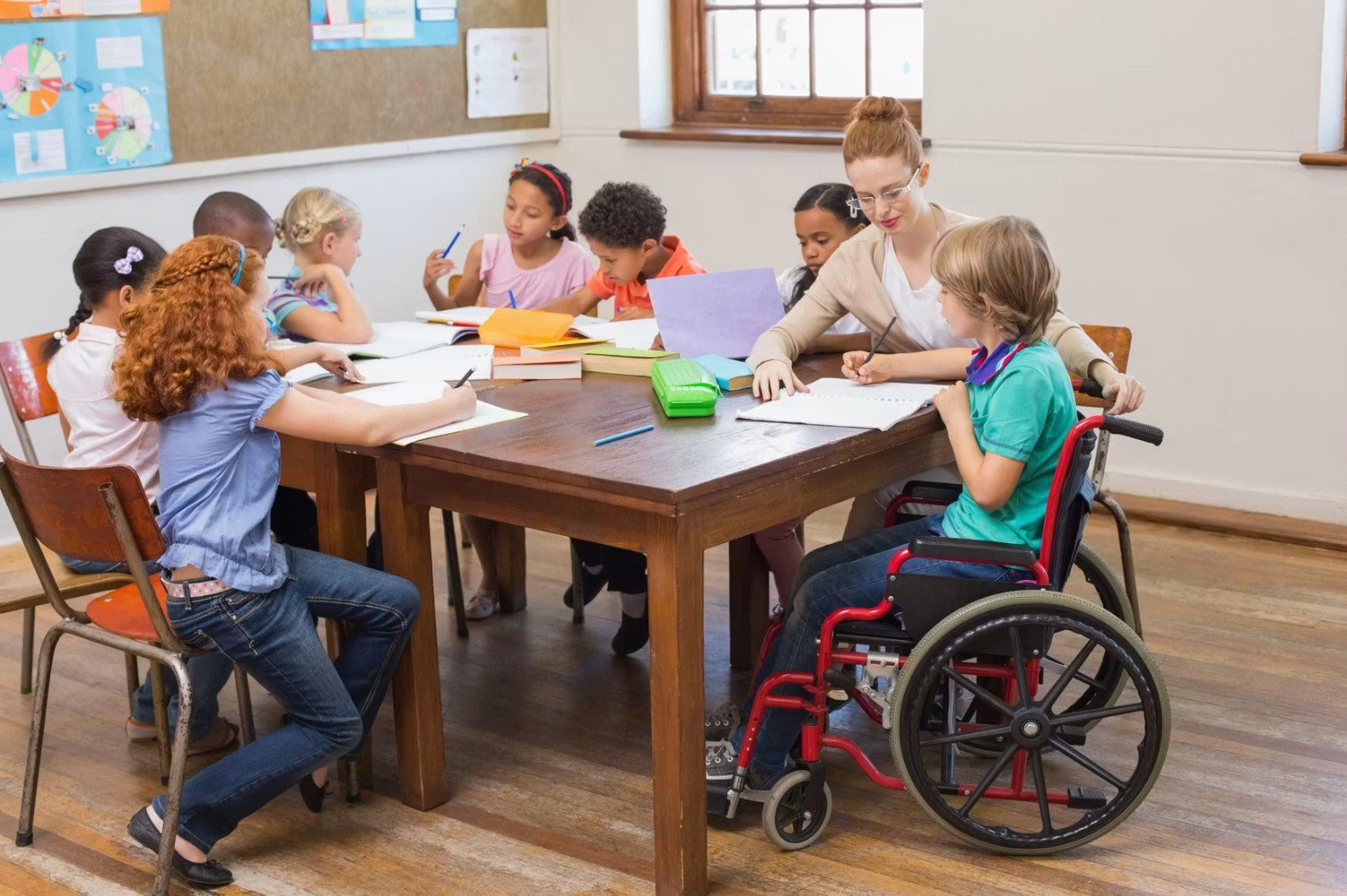 Children are shown seated around a table, talking and receiving instruction from a teacher. One child is seated in a red and black wheelchair, wearing a teal shirt.