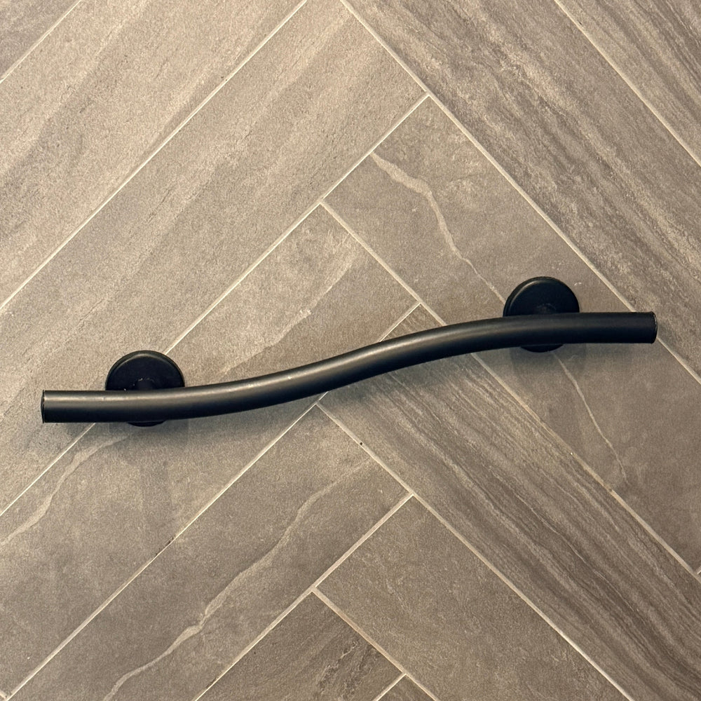 A curved matte black metal grab bar mounted on a gray herringbone tile wall inside a shower stall.