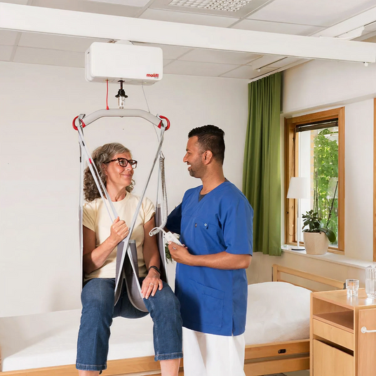 Person using a medical lift in a room with a window, bed, and side table, with a nurse assisting.