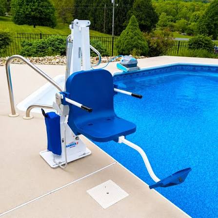 A blue and white pool chair lift mounted beside a swimming pool on a concrete deck, with a pool cleaner and landscaped greenery in the background.