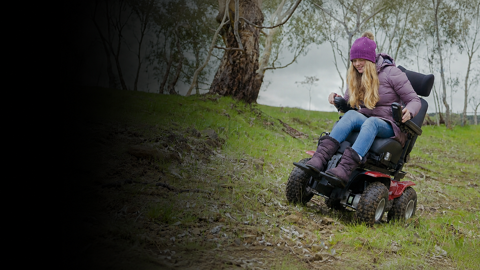 A woman wearing a purple beanie and a purple puffer jacket sits on a red and black motorized wheelchair on a grassy, wooded hillside.