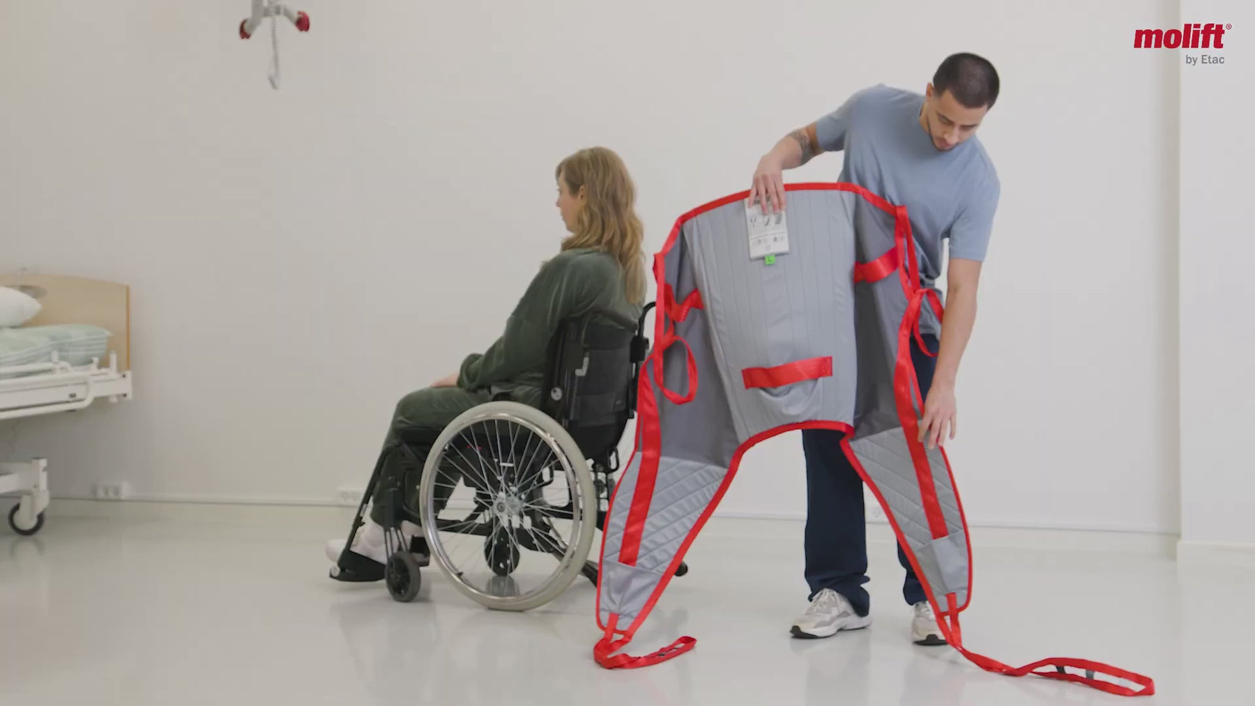 Instructional video demonstrating how to fold the Molift RgoSling Padded and correctly apply it to a person seated in a wheelchair, showing the sling positioned behind the back, threaded around the legs, and secured for safe use with a patient lift. No spoken narration; background music only.